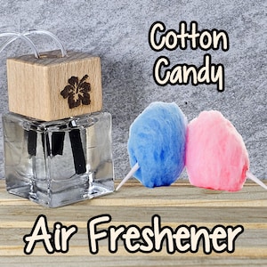 May include: A clear glass bottle with a wooden lid and a hibiscus flower design. The bottle contains a liquid air freshener with the scent of cotton candy. The bottle is sitting on a wooden surface with two cotton candy puffs in the background. The text "Cotton Candy" and "Air Freshener" is visible in the image.