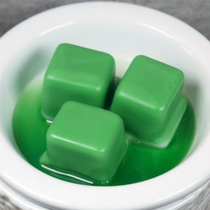 Puede incluir: Tres fundentes de cera verdes en un cuenco de cer&aacute;mica blanco. Los fundentes de cera son de forma cuadrada y est&aacute;n parcialmente derretidos.