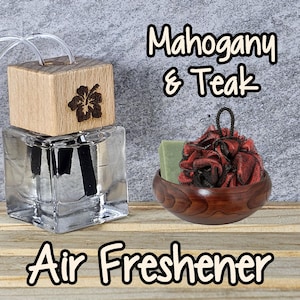 May include: A glass air freshener bottle with a wooden lid and a hibiscus flower design. The bottle is filled with a clear liquid and has two black reeds. The bottle is sitting on a wooden surface. The text "Mahogany & Teak Air Freshener" is visible in the image.
