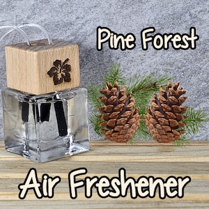 May include: A clear glass bottle with a wooden lid and a floral design. The bottle contains a liquid air freshener with a scent of pine forest. The bottle is sitting on a wooden surface with two pine cones and pine needles.