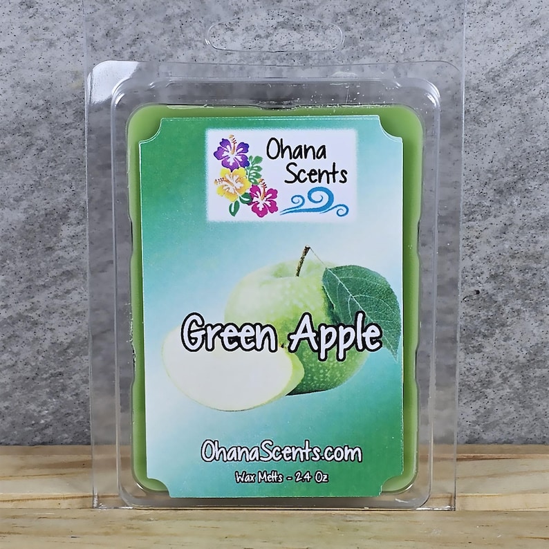 Puede incluir: Un fundente de cera con aroma a manzana verde en un envase de pl&aacute;stico transparente con forma de concha. El fundente de cera es verde y tiene una imagen de una manzana verde. La etiqueta dice "Ohana Scents" y "Green Apple". La etiqueta tambi&eacute;n dice "Wax Melts - 2.4 Oz".