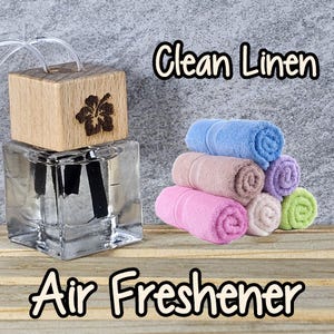 May include: A clear glass air freshener bottle with a wooden top featuring a flower design. Rolled, colorful towels in blue, pink, purple, green, and tan are stacked nearby. The text "Clean Linen" and "Air Freshener" are displayed.