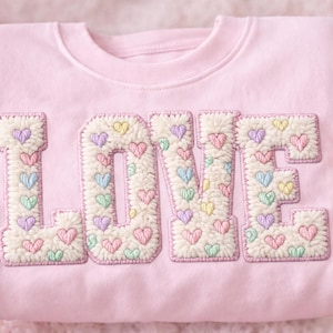 Könnte beinhalten: Hellrosa Sweatshirt mit dem Wort "LOVE" auf der Vorderseite gestickt. Die Buchstaben sind cremefarben mit einer flauschigen Textur und mit pastellfarbenen Herzen verziert. Das Sweatshirt wird von einer Person gehalten.