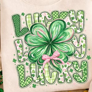 May include: Cream-colored sweatshirt with a large graphic that says "Lucky" in green letters. The "O" in lucky is replaced with a large green and pink shamrock with a pink bow. The letters have a variety of patterns.