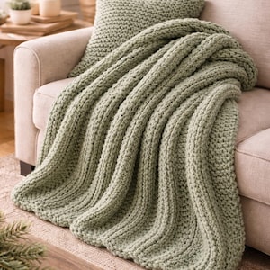 Puede incluir: Una manta y un cojín de punto verde salvia descansan sobre un sofá beige. La manta de punto grueso está drapeada sobre el sofá, creando un ambiente acogedor y acogedor. La manta y el cojín están hechos de un material suave y texturizado.