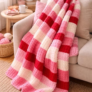 以下が含まれることがあります： 赤、ピンク、白のストライプ模様の手編みの毛布が、ベージュのソファに掛けられています。毛布の模様は、部屋に質感と暖かさを加えています。太い編み方は、快適さとリラックスを連想させます。
