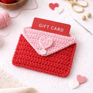 May include: A handmade red and pink crochet gift card holder shaped like an envelope. The flap is light pink with a heart accent and a button closure. A red gift card is visible inside. Other craft supplies are in the background.