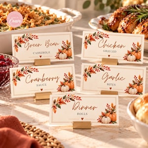 May include: Five rectangular food place cards with gold metal stands, featuring watercolor autumn leaf and pumpkin illustrations. The cards are labeled Green Bean Casserole, Grilled Chicken, Cranberry Sauce, Garlic Bites, and Dinner Rolls in brown text.
