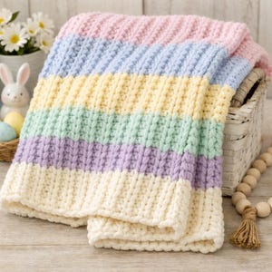 May include: A pastel-striped crochet blanket in shades of pink, blue, yellow, green, purple, and cream. The blanket is folded over a white wicker basket. Easter decorations, including a rabbit and eggs, are visible in the background.