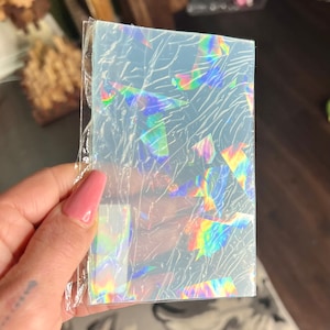 May include: A rectangular, iridescent sheet with a rainbow holographic effect, encased in clear plastic. The sheet displays a fractured pattern, reflecting light in multiple colors. The item is held in a hand.