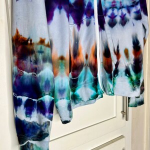 Pode incluir: Uma jaqueta de moletom azul, roxo e laranja tie-dye com um z&iacute;per branco. A jaqueta est&aacute; pendurada em uma porta de arm&aacute;rio branca.