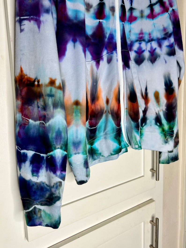 Pode incluir: Uma jaqueta de moletom azul, roxo e laranja tie-dye com um z&iacute;per branco. A jaqueta est&aacute; pendurada em uma porta de arm&aacute;rio branca.