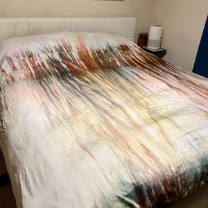 Könnte beinhalten: Eine weiße Bettdecke mit einem farbenfrohen abstrakten Design in Braun-, Orange- und Grüntönen. Die Bettwäsche befindet sich auf einem Bett mit einem weißen Kopfteil. Das Kunstwerk an der Wand über dem Bett hat ein farbenfrohes abstraktes Design.