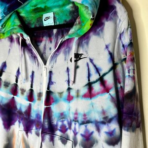 Pode incluir: Um moletom branco com z&iacute;per e um padr&atilde;o tie-dye em tons de roxo, azul, verde e laranja. O moletom tem um logotipo Nike preto no peito esquerdo e um capuz com cord&atilde;o preto.