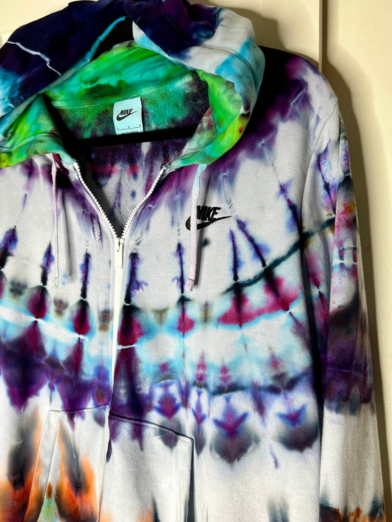 Pode incluir: Um moletom branco com z&iacute;per e um padr&atilde;o tie-dye em tons de roxo, azul, verde e laranja. O moletom tem um logotipo Nike preto no peito esquerdo e um capuz com cord&atilde;o preto.