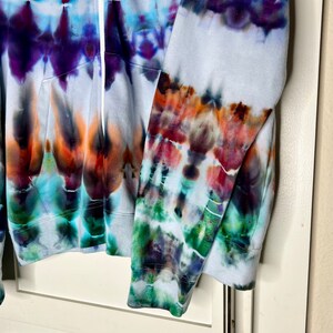 Pode incluir: Uma jaqueta de moletom branca com tie-dye e fecho de z&iacute;per. A jaqueta tem um padr&atilde;o de tie-dye colorido em tons de azul, roxo, laranja, verde e marrom.
