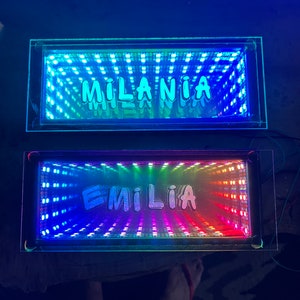 Peut inclure: Deux présentoirs lumineux à LED rectangulaires avec les noms "MILANIA" et "EMILIA" en lettres à bulles. Les lumières créent un effet miroir infini avec des teintes bleues, vertes et arc-en-ciel.