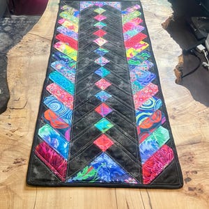 Op de afbeelding: Een rechthoekige tafelloper met een zwarte achtergrond en kleurrijke, patroonaccenten. De loper heeft een centraal ruitpatroon en randen met levendige bloemen- en abstracte ontwerpen. De loper ligt op een houten tafel.