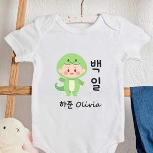 May include: White baby onesie with a green snake character wearing a hat. The onesie has the text "뱀" and "일" in Korean and "하준 Olivia" in English.