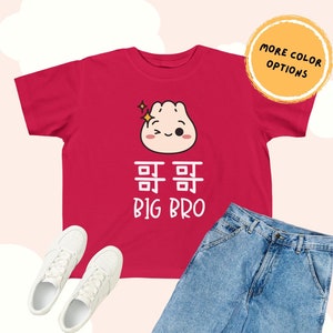 May include: A red t-shirt featuring a cartoon dumpling with a wink and sparkle. Below the graphic are the words "哥哥 BIG BRO" in white. The image also shows white sneakers and blue jeans.