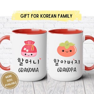 May include: Two white ceramic mugs with red interiors and handles. One mug features a pink pouch graphic with the word "Grandma" in English and Korean. The other mug has a persimmon graphic with the word "Grandpa". Text at the top says "Gift for Korean Family".