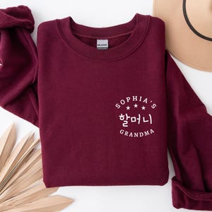 May include: A burgundy sweatshirt with the text "Sophia's Grandma" in white, including Korean characters. The sweatshirt has a crew neck and long sleeves with rolled-up cuffs. A tan hat is visible in the background.