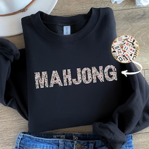 May include: A black sweatshirt featuring the word "MAHJONG" in a pattern of Mahjong tiles. The sweatshirt is folded, with blue jeans at the bottom. A white hat is visible in the background. The image includes a close-up of Mahjong tiles.