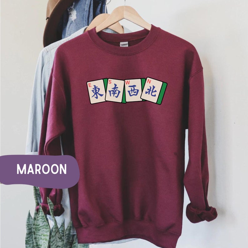 May include: Maroon sweatshirt with a crew neck. The front features a graphic of four Mahjong tiles with Chinese characters and the letters E, S, W, and N. The word "MAROON" is in a purple rectangle at the bottom.