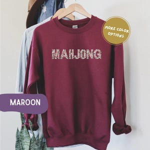May include: A maroon-coloured sweatshirt featuring the word "MAHJONG" in a patterned design. The long-sleeved top has a crew neck. Additional text includes "MORE COLOR OPTIONS" and "MAROON".