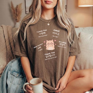 May include: A brown t-shirt with Korean text and a cat graphic. The shirt features phrases like "I Love You," "OMG!," "Older brother," "Thank You," and "It's okay." The cat graphic is pink and has hearts around it.