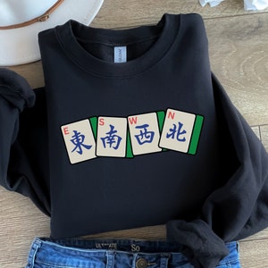 May include: Black sweatshirt featuring four Mahjong tiles with the characters for East, South, West, and North. The tiles are in a row, with green borders and white backgrounds. The sweatshirt is paired with blue jeans.