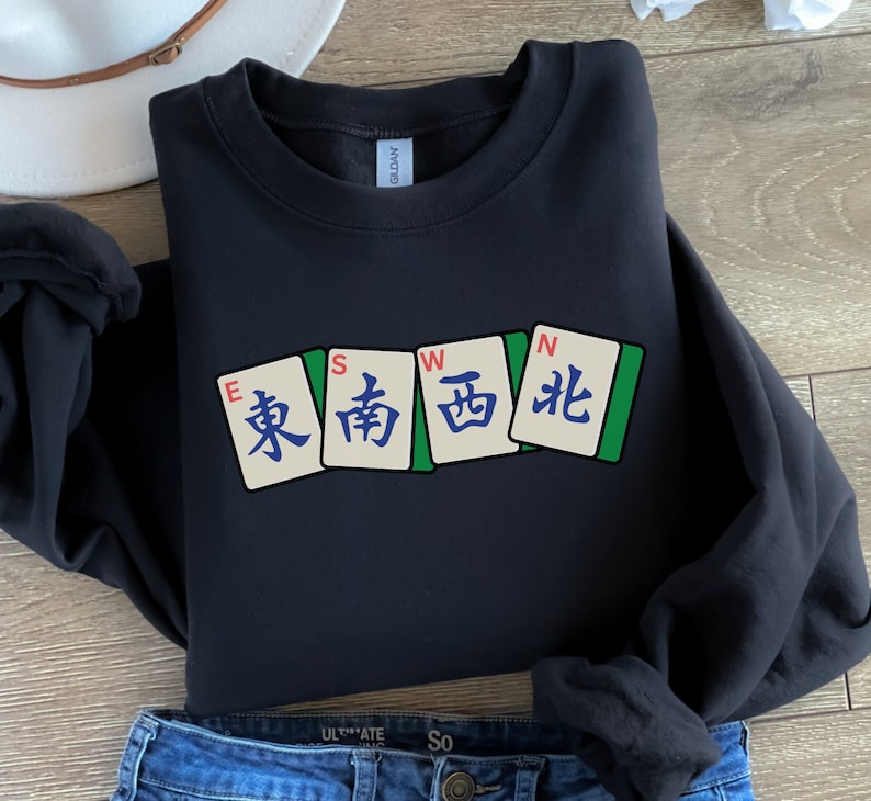 May include: Black sweatshirt featuring four Mahjong tiles with the characters for East, South, West, and North. The tiles are in a row, with green borders and white backgrounds. The sweatshirt is paired with blue jeans.