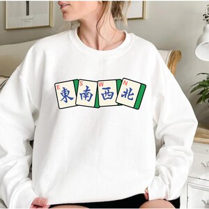May include: White sweatshirt featuring a graphic of four Mahjong tiles. The tiles display the characters for East, South, West, and North in blue and green. The sweatshirt is a casual, comfortable style.