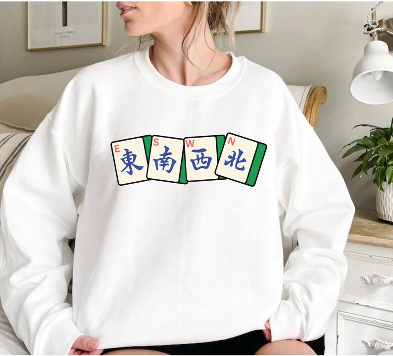 May include: White sweatshirt featuring a graphic of four Mahjong tiles. The tiles display the characters for East, South, West, and North in blue and green. The sweatshirt is a casual, comfortable style.