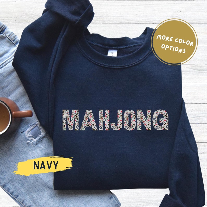 May include: Navy blue sweatshirt featuring the word "MAHJONG" in a patterned design. The sweatshirt has a crew neck and long sleeves. The text "MORE COLOR OPTIONS" is in a circle. The word "NAVY" is in a yellow rectangle.