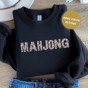May include: A black sweatshirt featuring the word "MAHJONG" in a design made of mahjong tiles. A gold circle with the text "MORE COLOR OPTIONS" is visible. The sweatshirt is styled with blue jeans and a white hat.