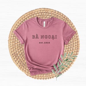May include: Mauve-colored t-shirt with rolled sleeves, featuring the text "BÀ NGOẠI" above "est.2026" in black. The shirt is displayed on a woven, circular placemat with a sprig of greenery.