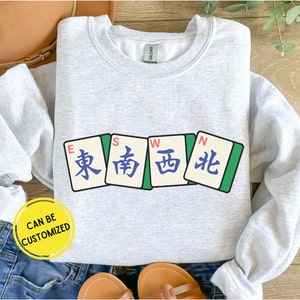 May include: Light gray sweatshirt featuring four Mahjong tiles with East, South, West, and North characters. The sweatshirt has the text "CAN BE CUSTOMIZED" in a yellow circle.