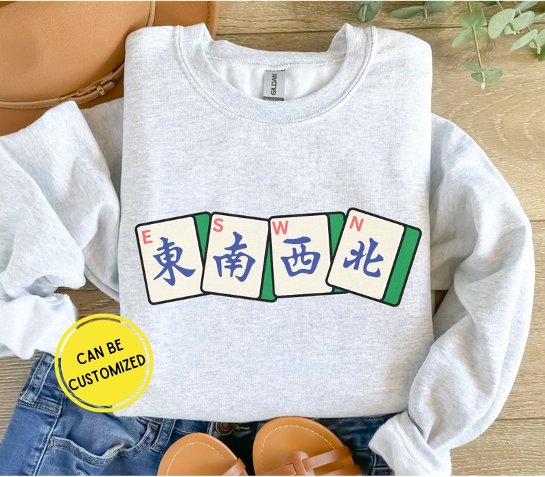 May include: Light gray sweatshirt featuring four Mahjong tiles with East, South, West, and North characters. The sweatshirt has the text "CAN BE CUSTOMIZED" in a yellow circle.