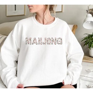 May include: White crew neck sweatshirt featuring the word "MAHJONG" in a pattern of Mahjong tiles. The long-sleeved top is made of a soft material. The design is casual and the text is the main focus of the garment.
