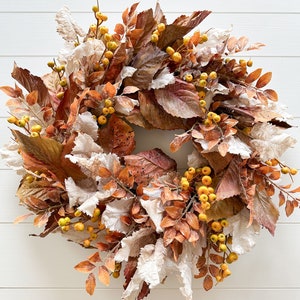 May include: A circular wreath composed of dried-look foliage in shades of brown, rust, and cream, interspersed with clusters of small, round, yellow berries. The arrangement features textured, crinkled leaves and delicate stems arranged in a dense, layered pattern.