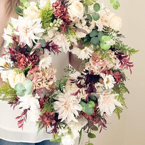 Peut inclure: Une couronne florale circulaire composée d'un mélange de roses crème, de dahlias blancs et d'hortensias rose poudré. L'arrangement est agrémenté de feuillage bordeaux profond, de feuilles d'eucalyptus vertes et de frondes de fougère délicates.