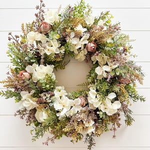 Peut inclure: Une couronne circulaire composée d'un arrangement dense de fleurs d'hortensia crème, de boutons de rose rose poudré et de divers feuillages verts, incluant de l'eucalyptus et des fougères, agrémentée de petits éléments botaniques séchés marron et beige.