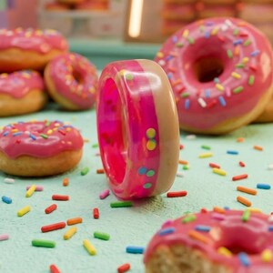 May include: A pink and tan donut-shaped ring with colorful sprinkles stands upright, surrounded by other donuts with pink frosting and sprinkles. The background is a light blue surface with scattered sprinkles.