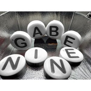 以下が含まれることがあります： 銀色の表面に「GABE NINE」という単語を綴る白い文字タイル