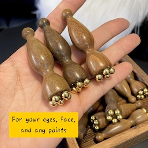 以下が含まれることがあります： 金色の金属製のボールがついた木製マッサージツールのセット。ツールは木製の箱に入っています。黄色い背景に「目、顔、そしてあらゆるポイントのために」と書かれています。