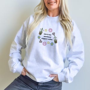 May include: A light gray sweatshirt with the text "this is my emotional support coffee" in black, along with cartoon images of coffee drinks, stars, flowers, and smiley faces. The sweatshirt is worn with ripped black jeans and black and white sneakers.
