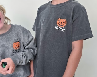 Puede incluir: Dos niños con camisetas a juego con temática de Halloween. Uno lleva una sudadera gris y el otro una camiseta gris. Ambas camisetas tienen un gráfico de calabaza naranja y un nombre. El niño de la izquierda lleva pantalones cortos color óxido.