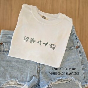 Peut inclure: Un t-shirt blanc à col rond orné d'outils de jardinage brodés en fil vert sauge argenté. Il est associé à un short en jean délavé clair avec des détails déchirés. Le texte indique que le t-shirt est blanc et le fil est vert sauge.