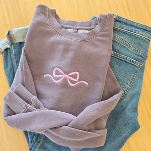 May include: Plum-colored sweatshirt with a lavender bow embroidery, paired with blue jeans. The sweatshirt's color is plum, and the thread color is lavender. The jeans are folded neatly next to the sweatshirt.
